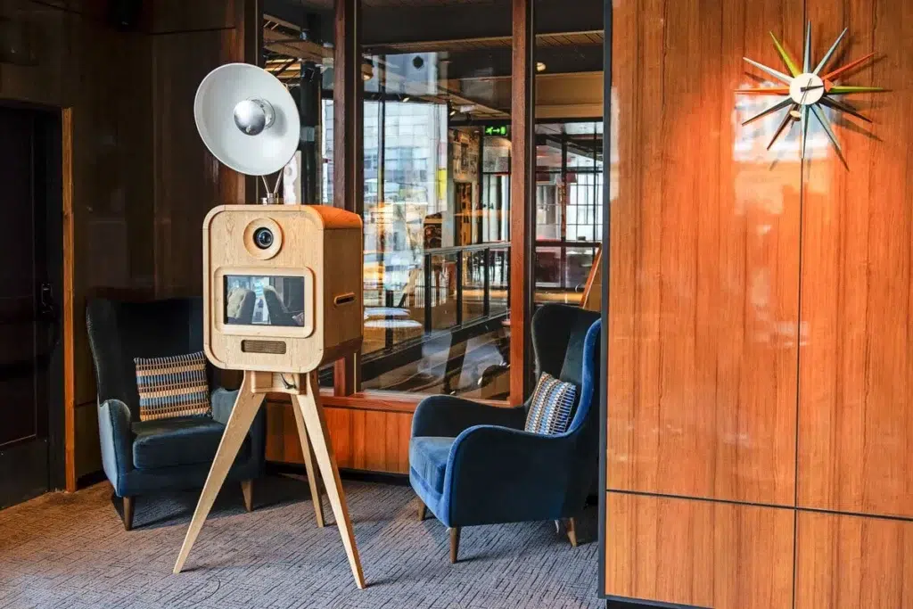 A modern photo booth with a wooden exterior stands between two blue velvet armchairs in a stylish room. A colourful wall clock hangs on wood-panelled walls, and large windows reflect the interior—perfect for sketch bot hire events.