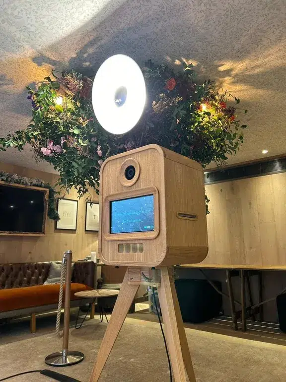 A wooden photo booth with a ring light stands on a tripod in a cosy room. Greenery and flowers create a decorative backdrop. Warm lighting, comfortable seating, and options like sketch bot hire add to the inviting atmosphere.