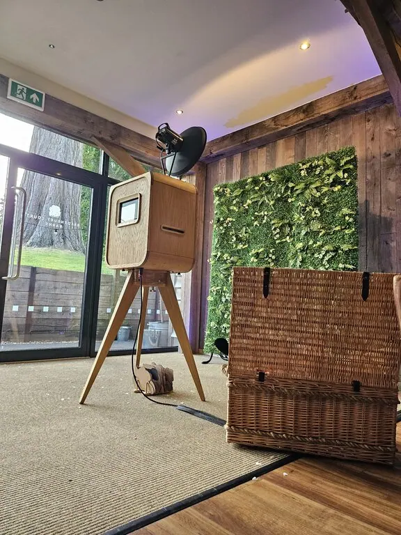 A premium photo booth hire with a camera and flash sits on tripod legs near a wicker basket, in a rustic room with wooden beams and a green foliage wall. A glass door lets in natural light from outside.