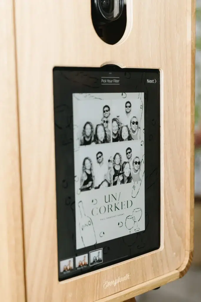 A wooden photo booth screen displays a collage of black and white photo strips featuring smiling people. The event name "UN/CORKED" appears at the bottom, along with wine-themed graphics—perfect for a memorable corporate event experiential activation.