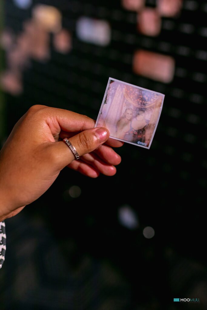 A hand with a silver ring holds a small, blurry photo of two people posing together. The dark, out-of-focus background features a wall with various other photos—perfect inspiration for graffiti wall hire at your next corporate event experiential.