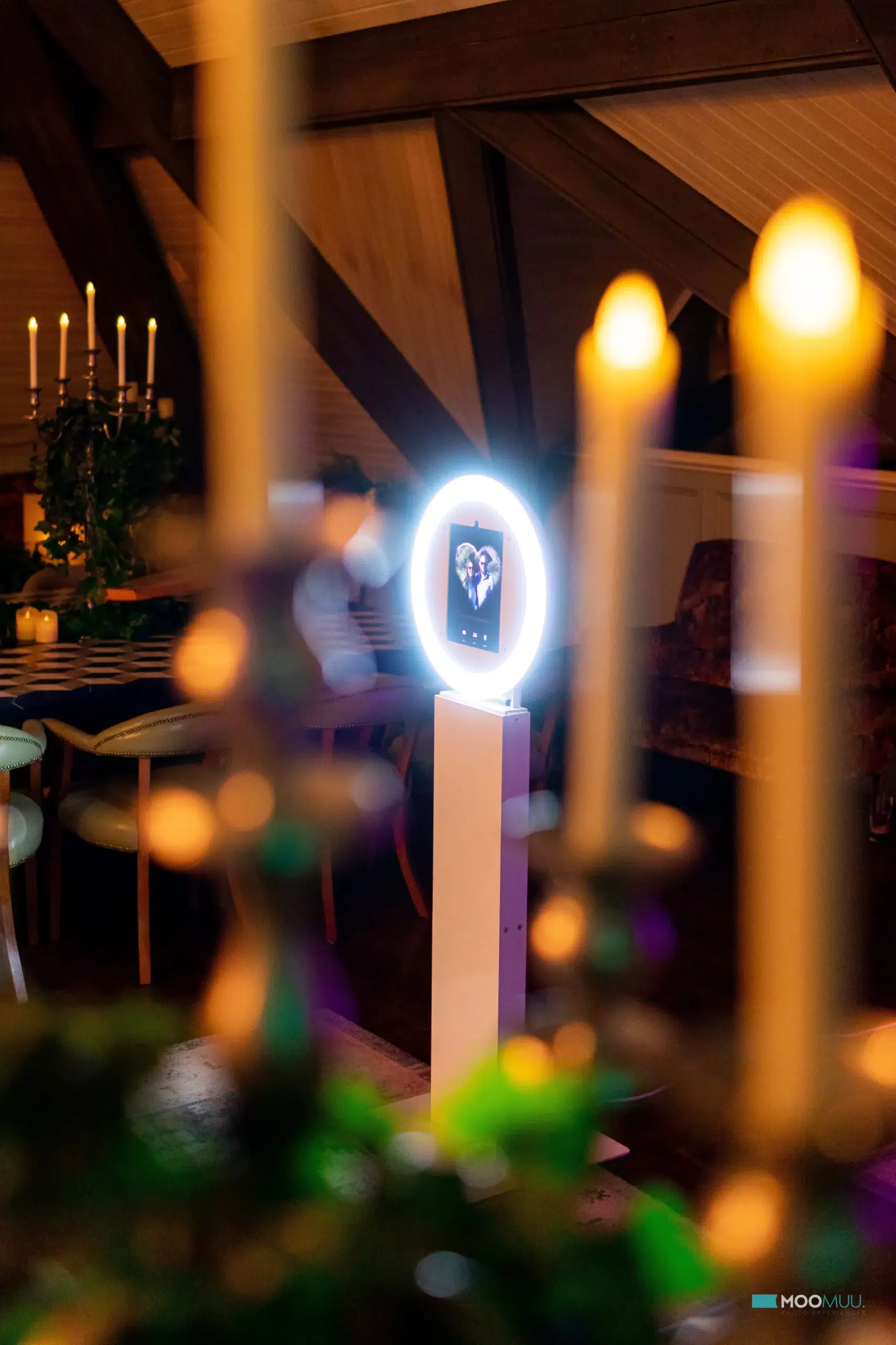 A warmly lit room features candles in the foreground and a premium photo booth hire with a circular LED light and screen at its centre. Cosy furniture and a candelabra decorate the background.