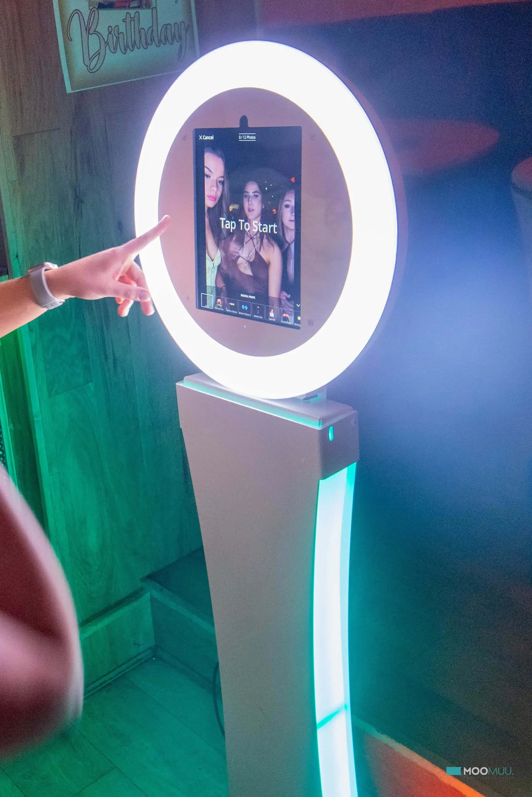 A person’s finger points at a photo booth touchscreen with a glowing ring light. The screen shows three people posing for a selfie, with the message “Tap To Start”—perfect for corporate event experiential experiences or sketch bot hire.