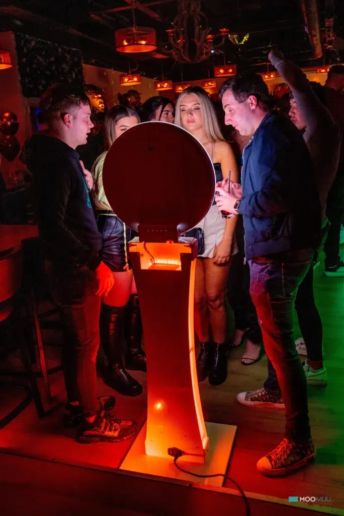 A group of people in a dimly lit bar gather round a photo booth, smiling and laughing as colourful lights illuminate their faces. Drinks and balloons set the scene for a fun corporate event experiential with sketch bot hire adding to the excitement.