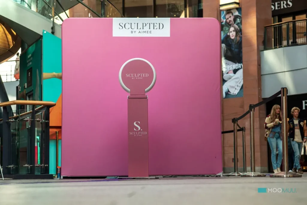 A pink promotional display for “Sculpted by Aimee” stands in a shopping centre, ideal for corporate event planning. It features a rectangular backdrop and pedestal with circular frame, whilst people and shops are visible in the background.