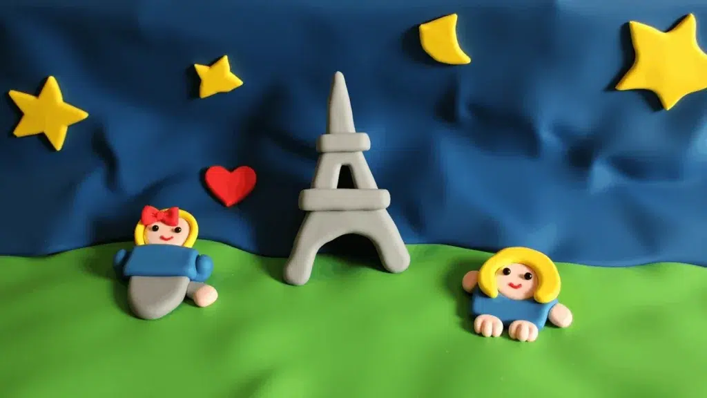 Clay figures of two smiling girls sit on green grass with a grey Eiffel Tower between them. Yellow stars, a crescent moon, and a red heart decorate the dark blue sky. Perfect for corporate event experiential displays or sketch bot hire inspiration.