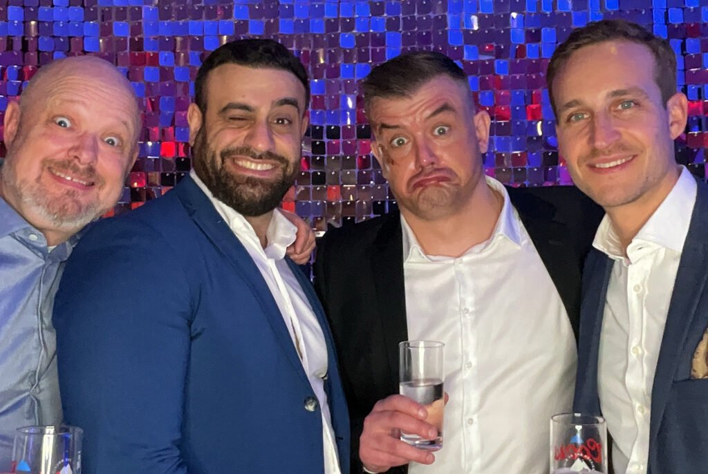 Four men in suits stand together in front of a sparkly purple backdrop, smiling and pulling funny faces whilst holding drinks—capturing the fun of unique AI photo experiences.