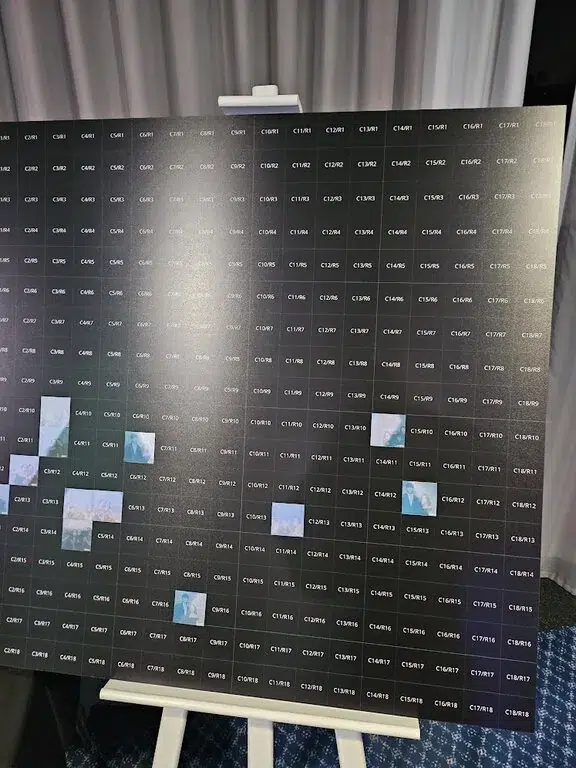 A large display board with small black squares labelled with codes, some featuring clouds or sky scenes. Perfect for a corporate event experiential, the board is set on an easel above a blue patterned carpet.
