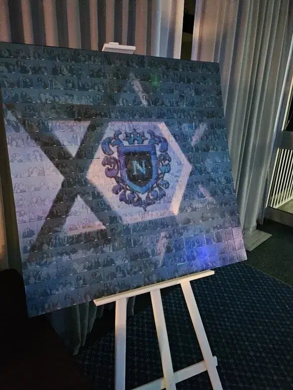 A large board on an easel displays a collage of small, faded photos forming a hexagon with a blue crest in the centre, set against a curtain and carpeted background—perfect for showcasing premium photo booth hire at your next corporate event experiential.