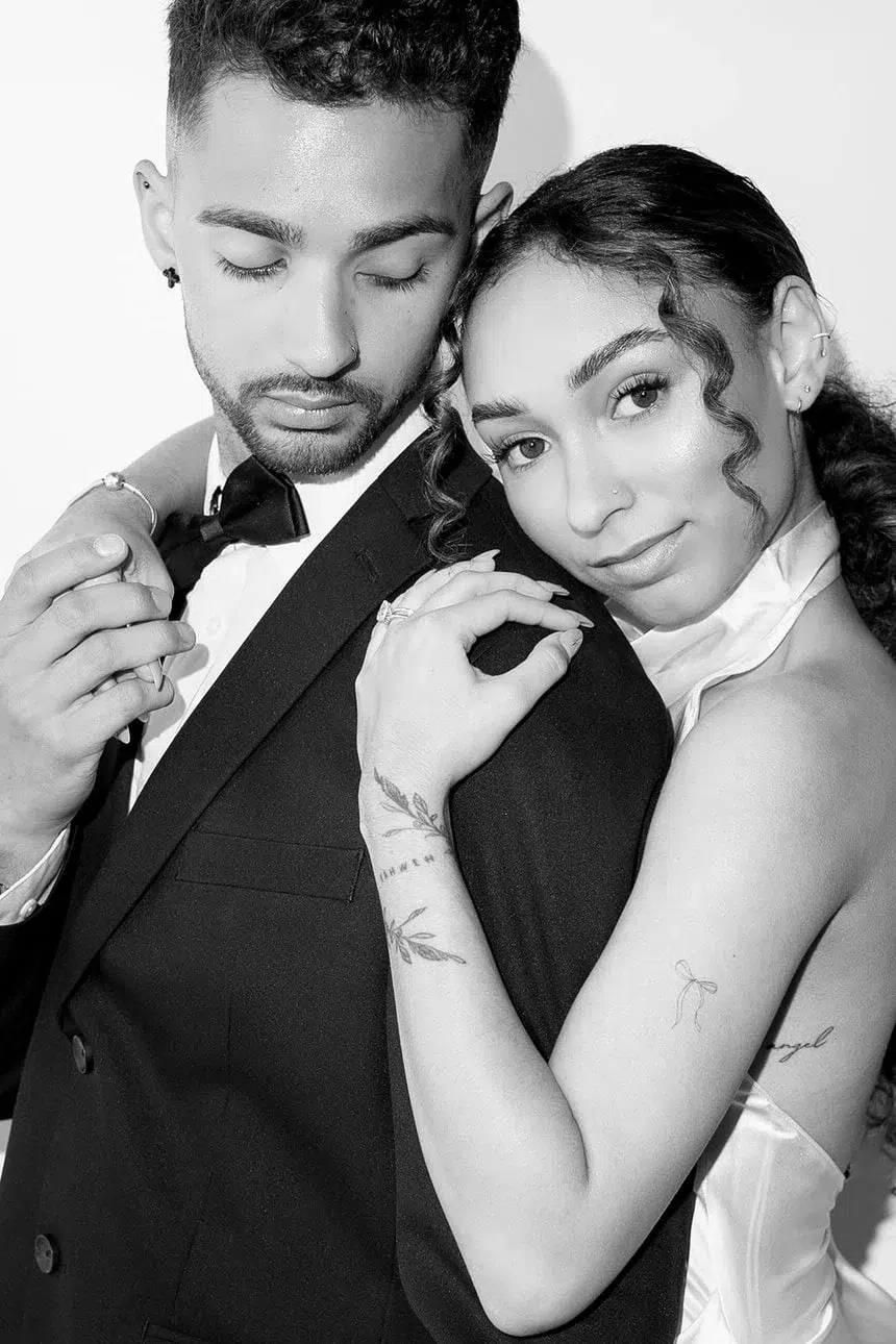 A woman in a halter dress embraces a man in a suit and bow tie from behind, posing before a vibrant mosaic wall. She smiles at the camera whilst he looks down, eyes closed. Both have visible tattoos and appear intimate and stylish.