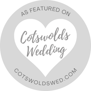 A circular grey badge with a white heart in the centre. Inside the heart, it says "Cotswolds Wedding." Around the edge, it reads "As featured on cotswoldswed.com"—perfect for showcasing Draw Me Bot or Photo Booth hire services.