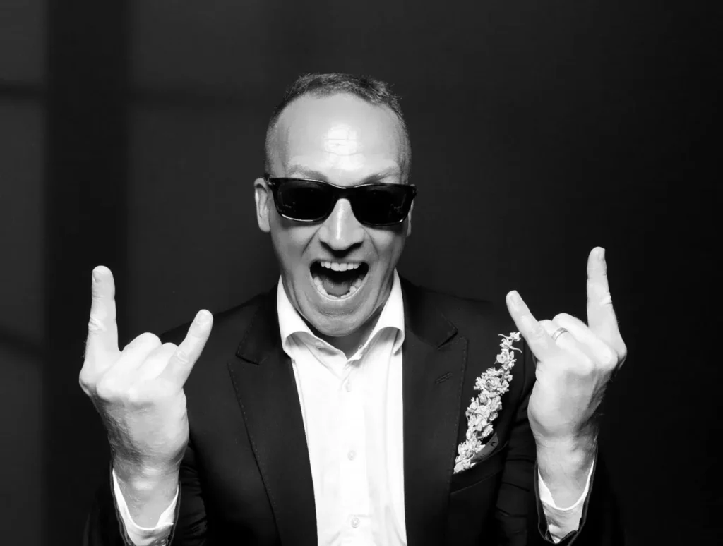 A man in a suit and sunglasses smiles widely, raising both hands in a "rock on" gesture against a dark background—perfect for capturing moments at a corporate event experiential or with premium photo booth hire.