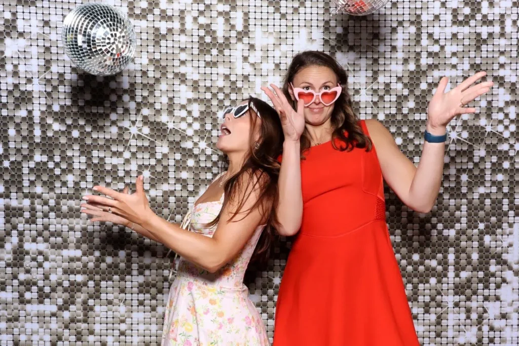 Two women wearing fun heart-shaped sunglasses pose and laugh in front of a sparkly silver sequin backdrop with disco balls, enjoying their time at a Photo Booth hire. One wears a floral dress, the other stuns in red.