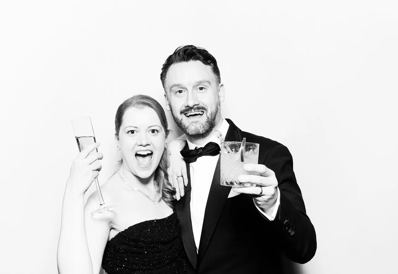 A smiling woman in a strapless dress and a man in a tuxedo pose together, each holding a drink and looking at the camera, captured with premium photo booth hire against a plain white background.