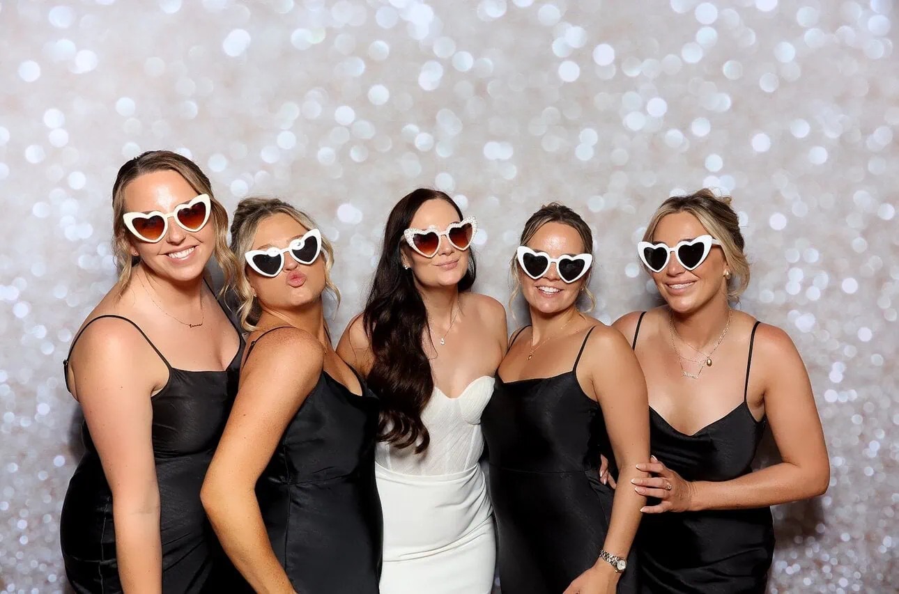 Five women, four in black dresses and one in white, pose together wearing white heart-shaped sunglasses against a sparkling, blurred background. Perfect for a fun group photo with premium photo booth hire. They are smiling and standing close to each other.