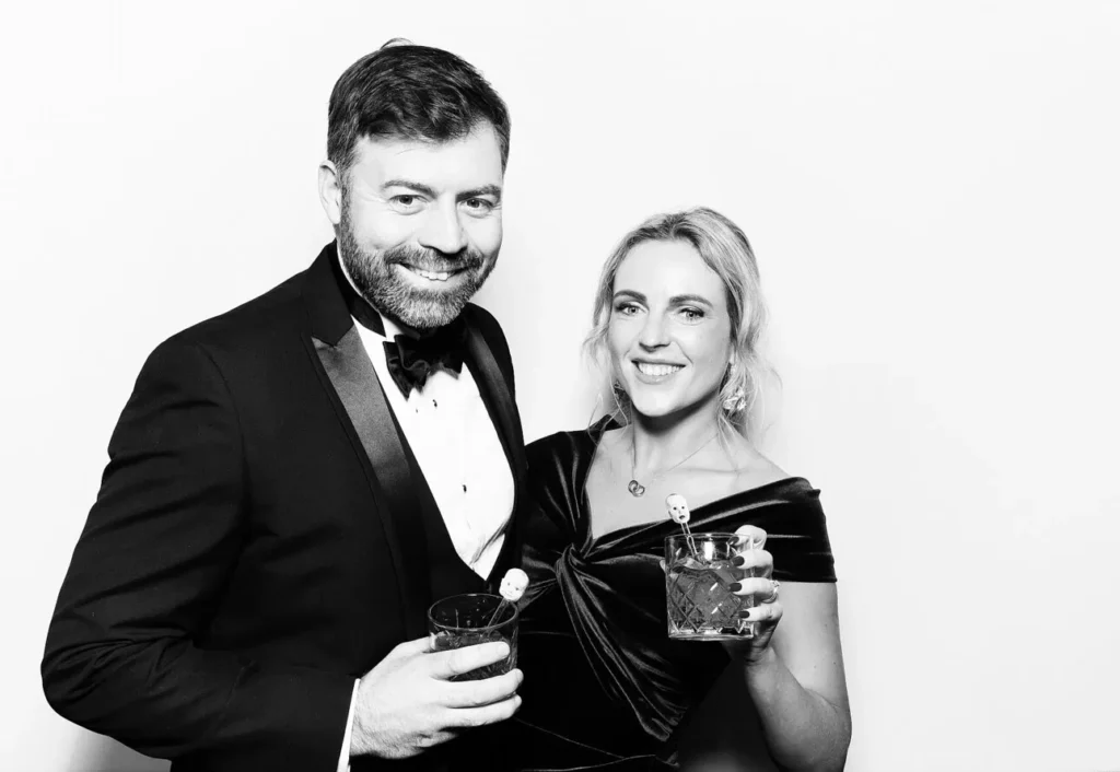 A man in a dinner jacket and a woman in an off-the-shoulder dress smile at the camera, each holding a drink with a decorative stirrer, in front of a plain white background—perfect for your next sketch bot hire or graffiti wall hire event.