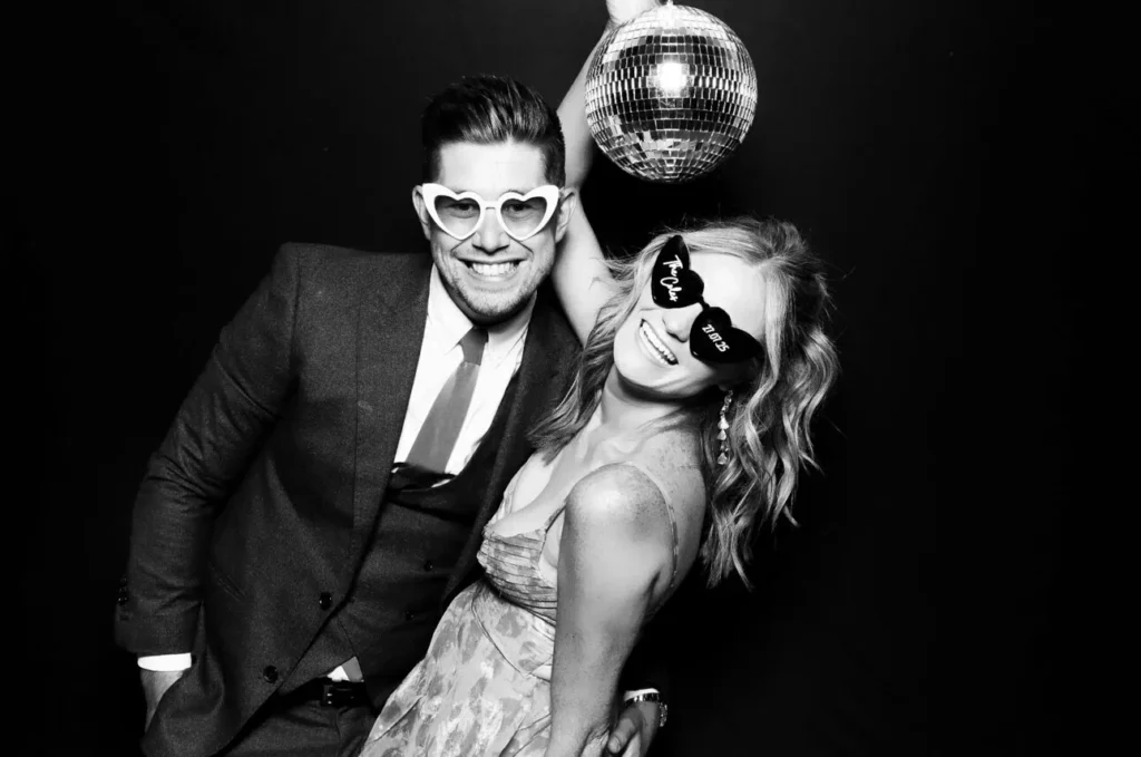 A man in a suit and a woman in a dress pose playfully with heart-shaped sunglasses. The woman holds a disco ball above them against a black background. Capture fun moments like this with Sketch Bot Hire for your next event!.