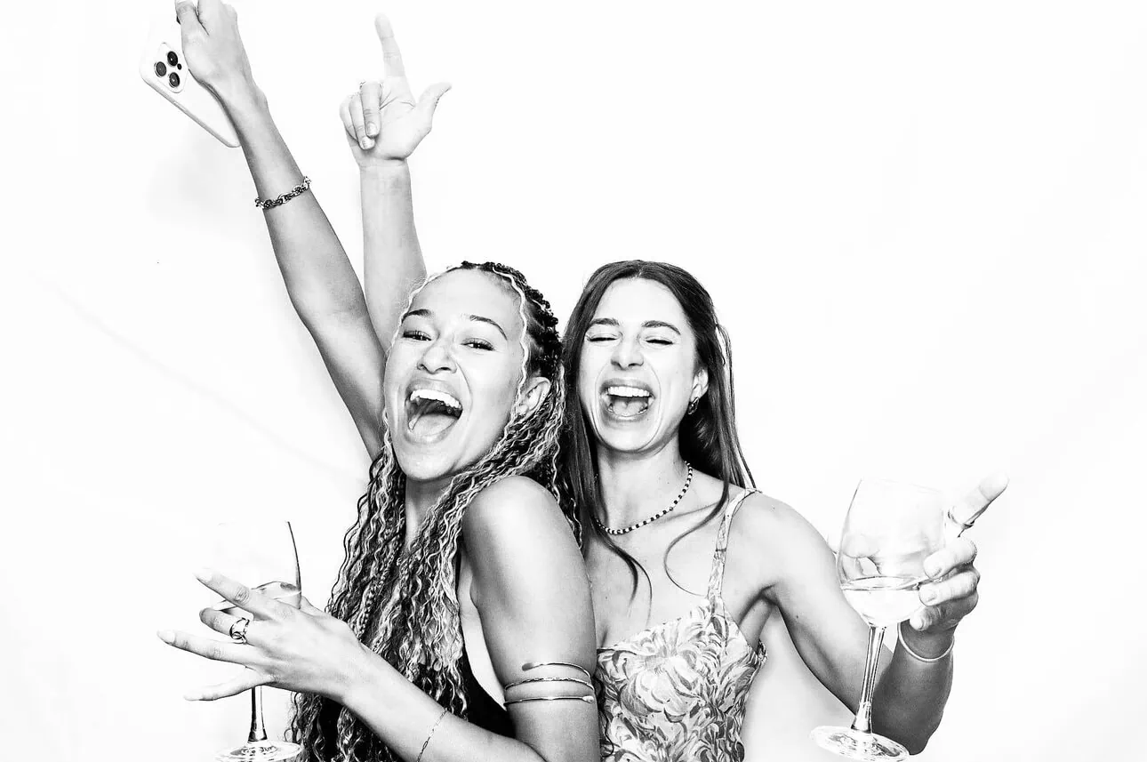 Two women smiling and laughing, holding wine glasses and posing energetically in front of a white background. One raises her mobile, pointing upwards, as they celebrate and have fun—perfect for a Draw Me Bot or Photo Booth hire experience!.