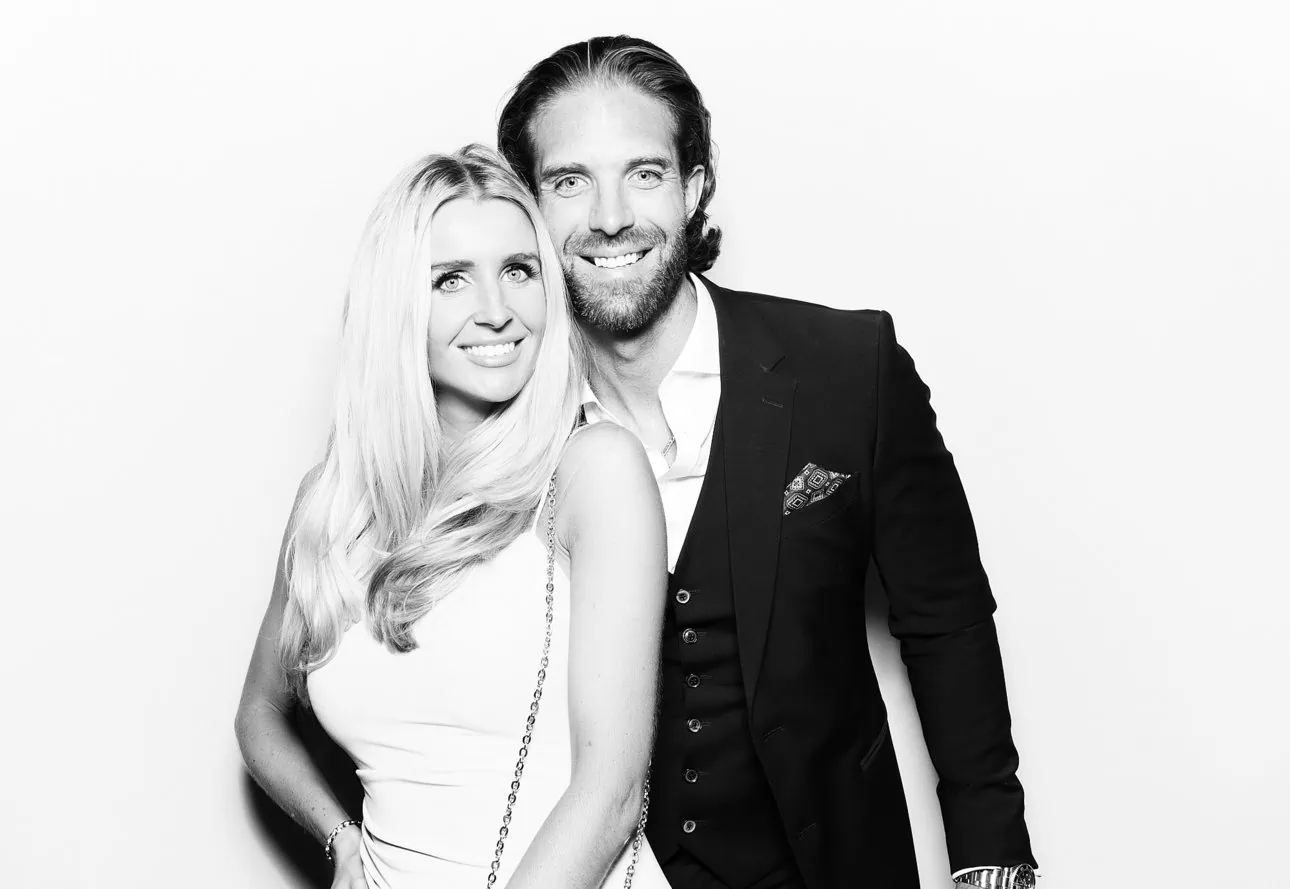 A smiling man in a suit and a smiling woman in a sleeveless dress stand close together against a plain, light background in this black and white photo—perfect for Photo Booth hire events.