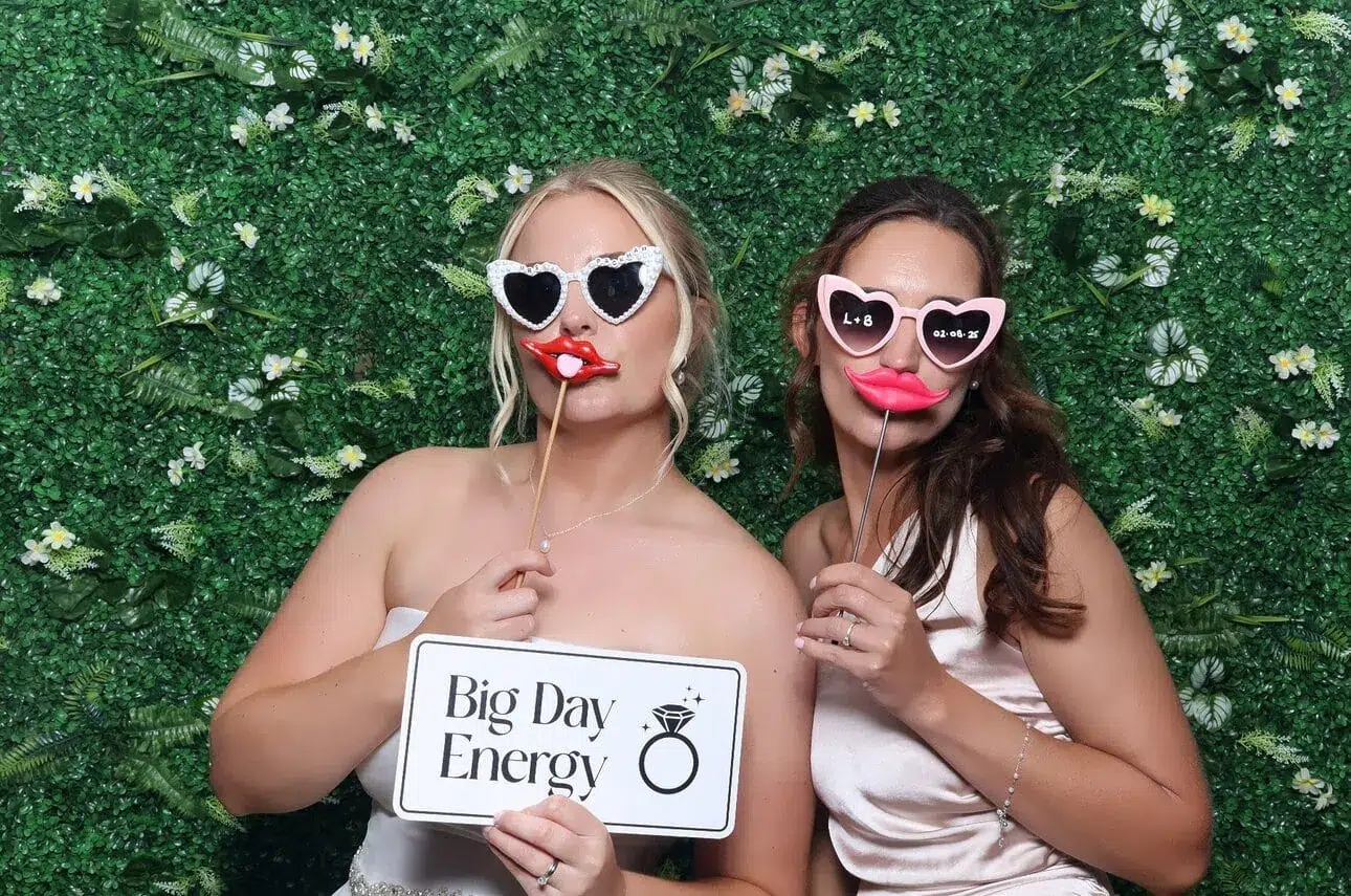green flower wall backdrop with a bride and her friend holding up a word sign and lips on sticks