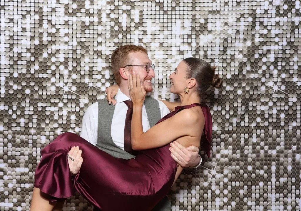 A man in a suit holds a woman in a burgundy dress as they smile at each other before a sparkling, silver sequin backdrop—a joyful moment perfectly captured at their Photo Booth hire for a formal event.