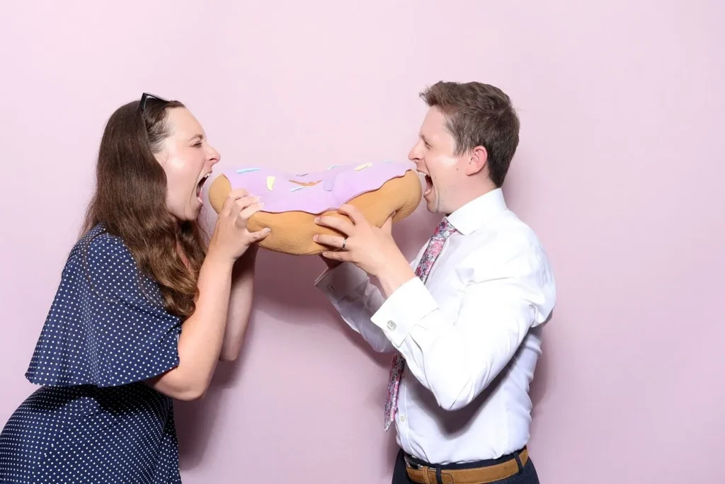 2 people standing in front of a pink backdrop holding a giant plush donut pretending to take a bite