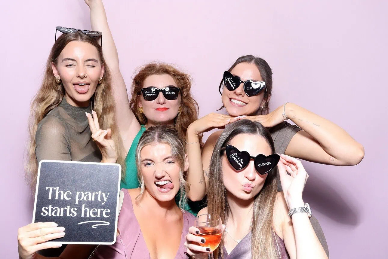 Five women pose playfully in front of a light pink backdrop. Some wear novelty sunglasses, hold drinks, and a sign reading "The party starts here." With smiles and silly faces, they're ready for memories—perfect for a Draw Me Bot or Sketch Bot Hire event!.
