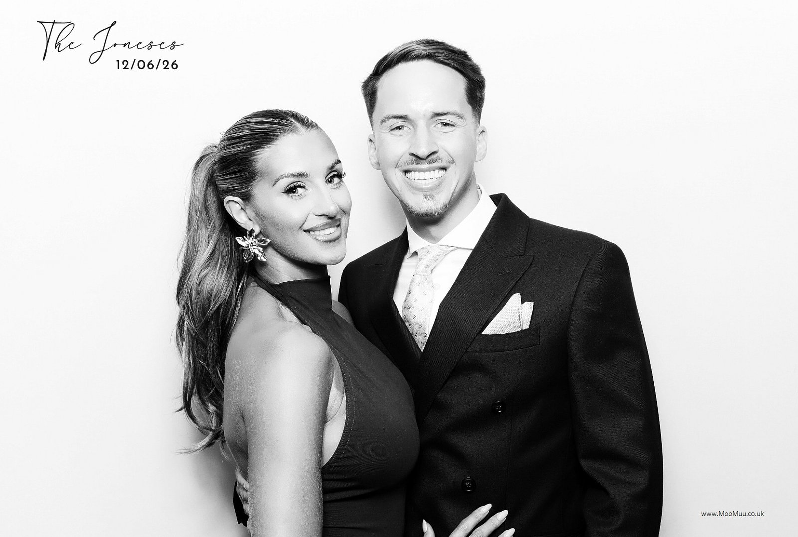 A smiling couple in formal attire poses in front of a white backdrop, capturing a memorable moment at an experiential photo booth. The woman wears a sleeveless dress with statement earrings, and the man sports a suit and tie. Text above reads "The Inverso 12/06/26".