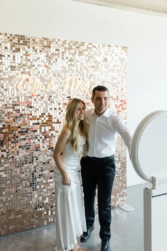 A smiling couple, dressed in formal attire, pose together in front of a sequinned "Mr & Mrs" backdrop while enjoying premium photo booth hire at an indoor event.