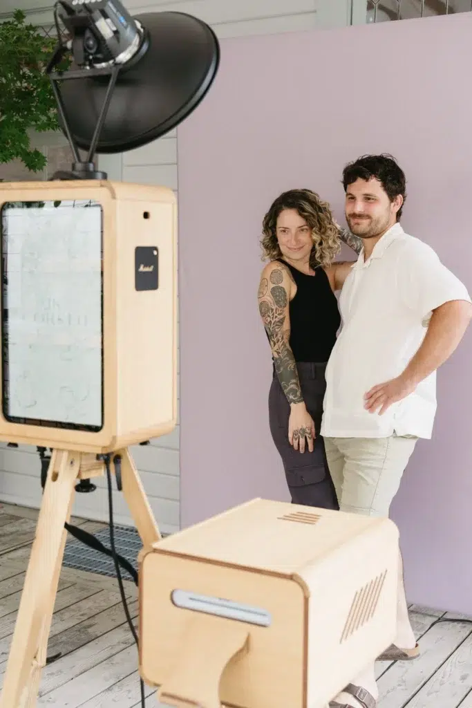A smiling couple pose together in front of a light purple backdrop, standing close with arms round each other, next to a wooden photo booth set-up with studio lighting—perfect for a corporate event experiential or graffiti wall hire.