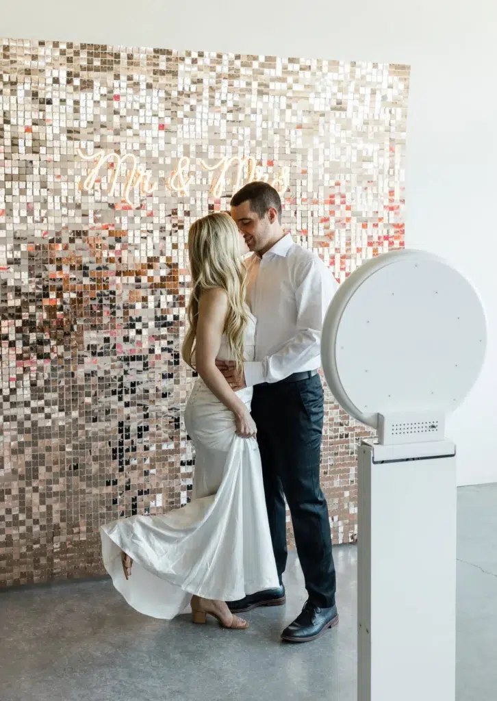 A bride and groom stand close together in front of a sparkling sequin wall with "Mr & Mrs" in neon script. The bride holds her white dress whilst the groom embraces her. A premium photo booth hire setup is in the foreground.