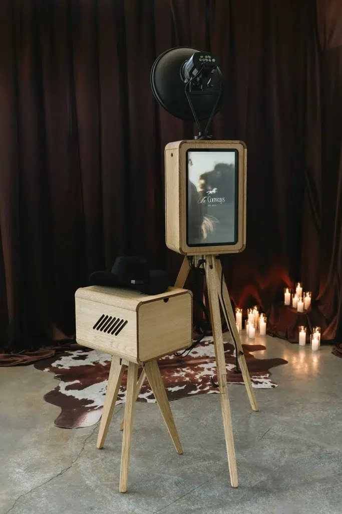 A modern wooden photo booth with a camera and ring light stands on a tripod beside a small matching table with a black hat on top. Candles and a cowhide rug decorate the background—perfect for premium photo booth hire.