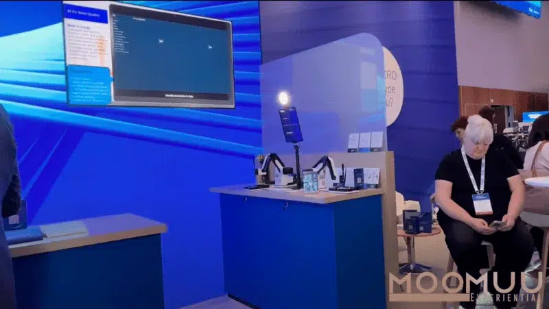 A person sits on a chair using their phone beside a blue trade show stand with digital screens, product displays, and a counter under bright lighting. The stand displays the "MOOMU Experiential" logo featuring MooMuu Edit.