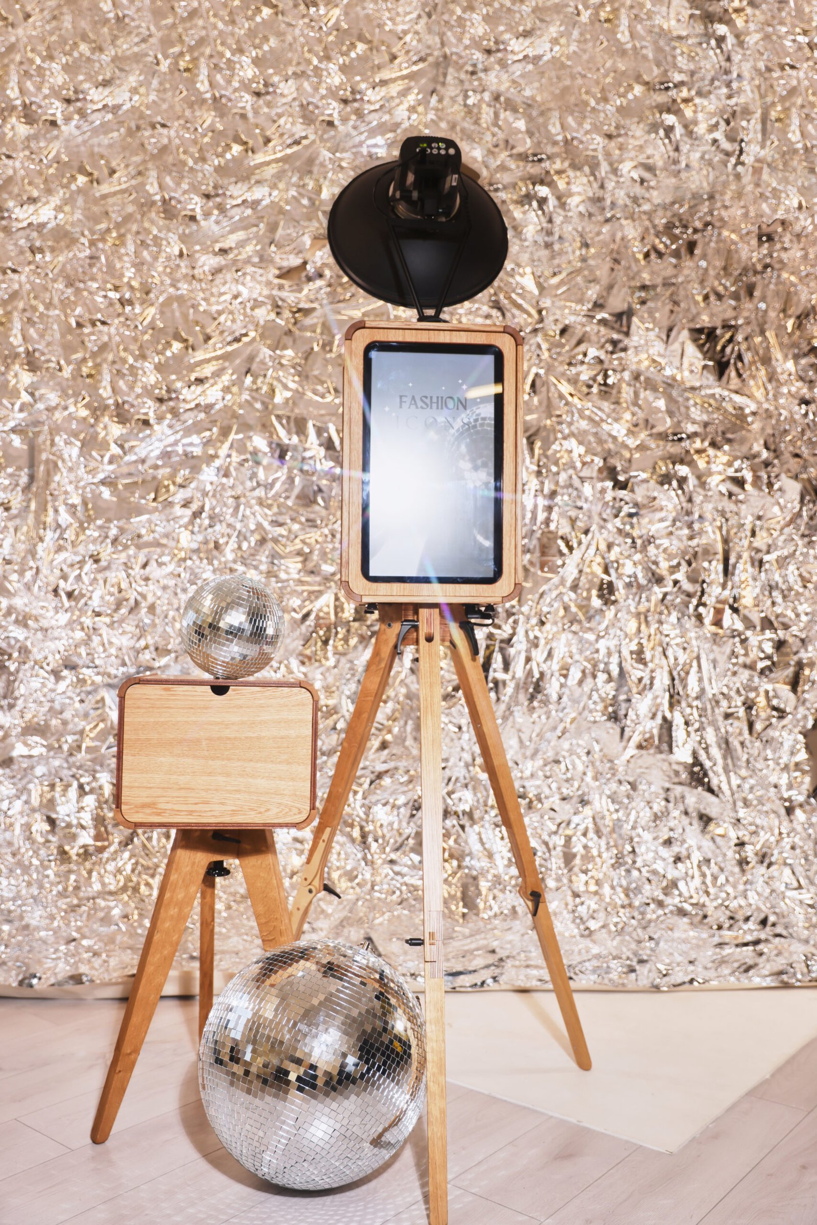 A wooden photo booth with a digital screen stands on a tripod in front of a shiny, silver tinsel backdrop. Beside it are two disco balls, one on a wooden stand and one on the floor—ideal for premium photo booth hire experiences.