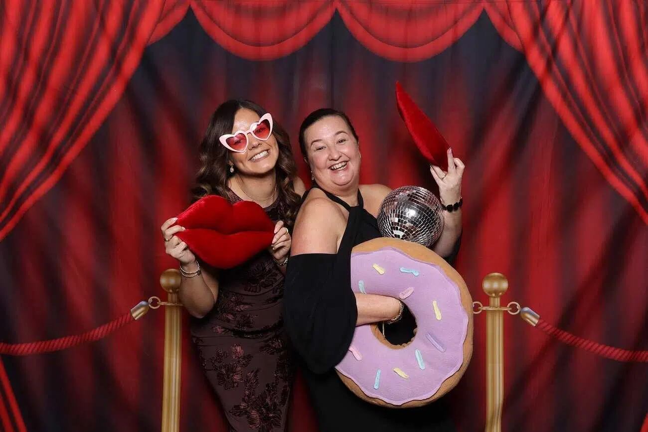 Two women pose in front of a red curtain backdrop, smiling. One wears heart-shaped glasses and holds a large plush red lips, while the other holds a disco ball and a plush doughnut—perfectly capturing the joy of celebrations.