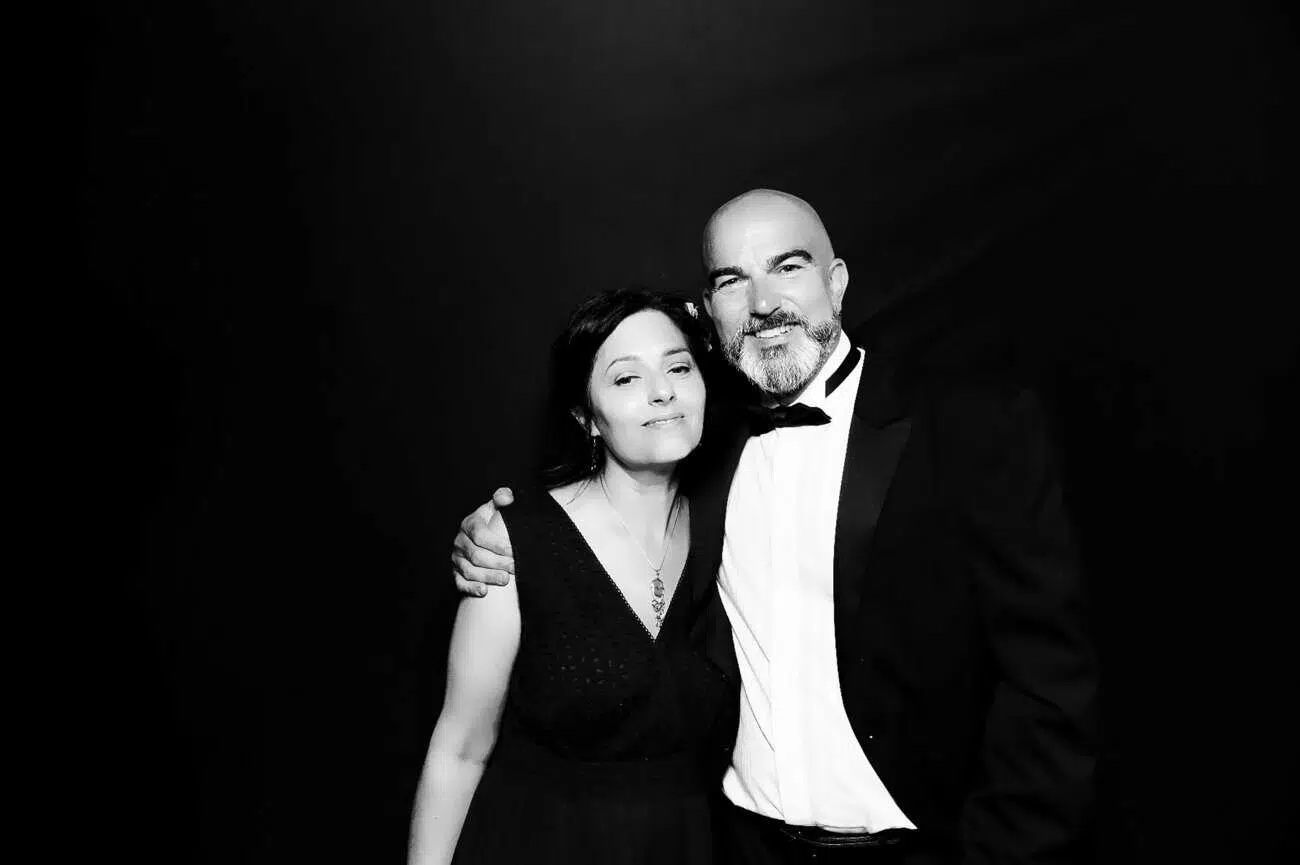A man in a dinner jacket and a woman in a sleeveless dress stand together, smiling with the man's arm around her shoulder, capturing the joy of celebrations against a plain dark background. The image is in black and white.