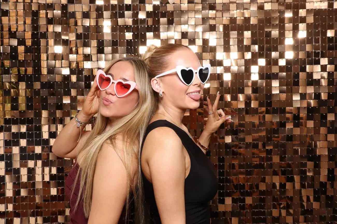 Two women pose playfully in front of a shiny gold sequin backdrop during celebrations, both wearing heart-shaped sunglasses and making peace signs, with one sticking out her tongue.