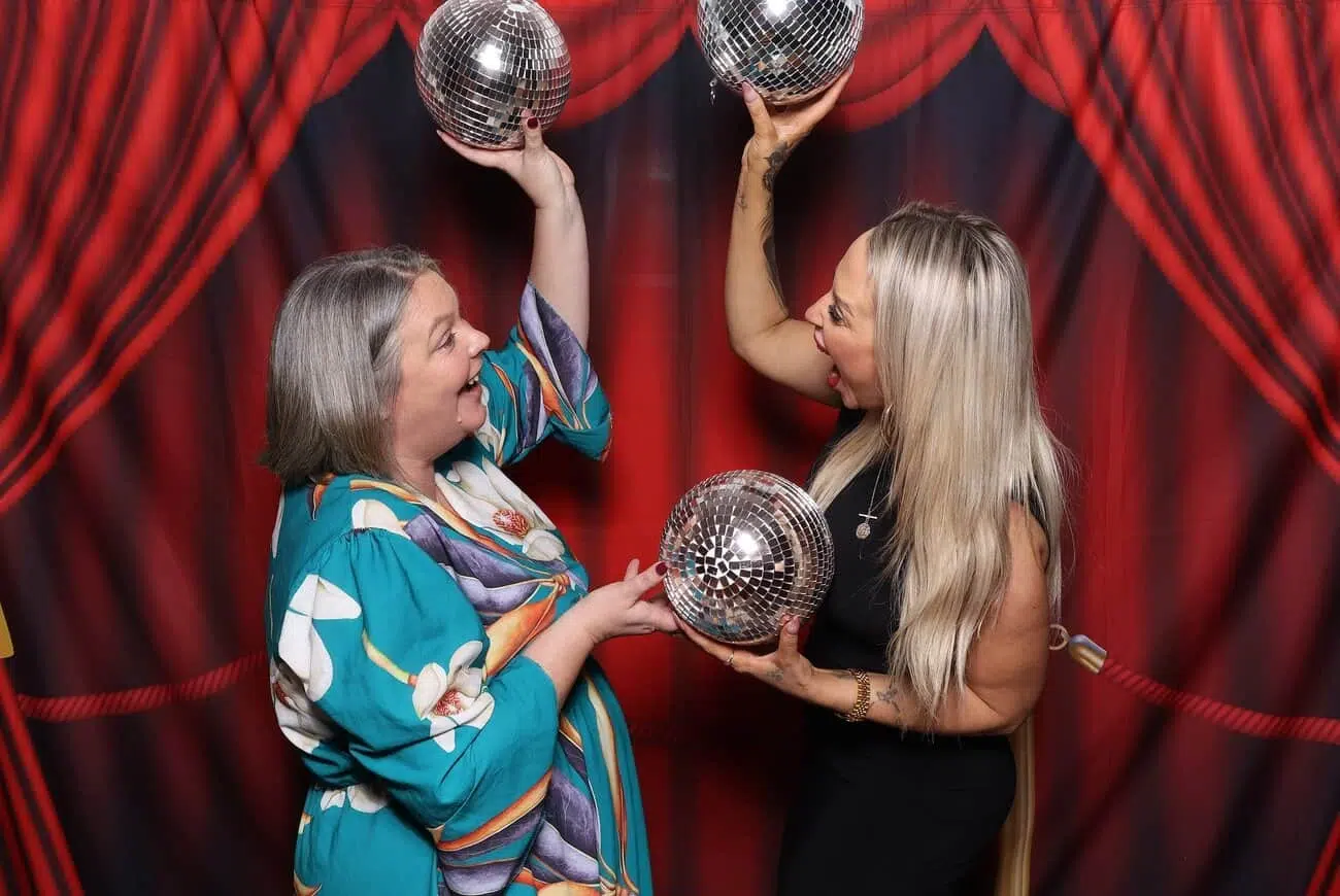 Two women stand in front of a red curtain backdrop, smiling and holding shiny disco balls above their heads, exuding the joyful spirit of celebrations.