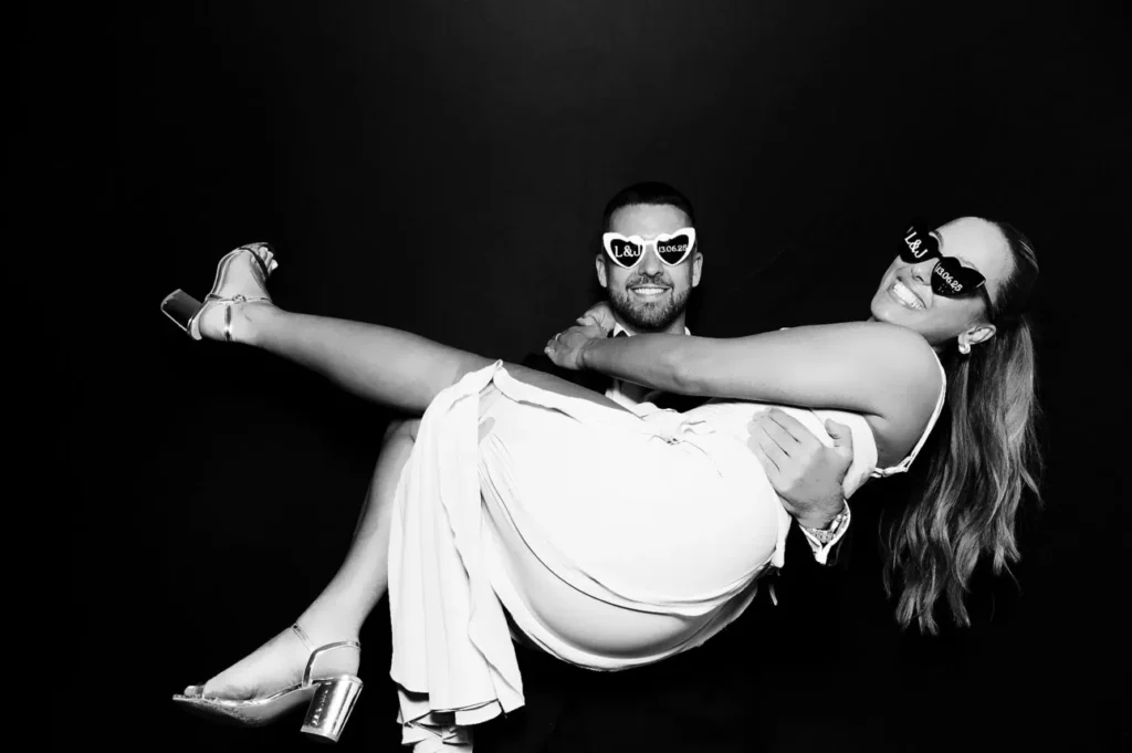 A man wearing sunglasses holds a smiling woman, both posing playfully in front of a plain dark background. This joyful moment could inspire premium photo booth hire for your next corporate event experiential.