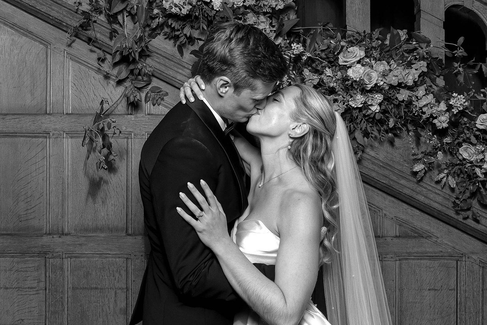 A bride and groom share a kiss in front of a flower-adorned wooden staircase. The moment is intimate and romantic—perfect for capturing with experiential photo marketing, creating unforgettable memories on their special day.