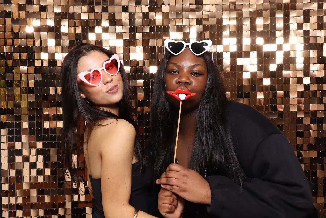 Two women pose in front of a sparkly gold sequin backdrop, celebrating with heart-shaped sunglasses. One holds playful red lips on a stick. Both smile in dark outfits, capturing the joy of celebrations in this fun photo booth moment.