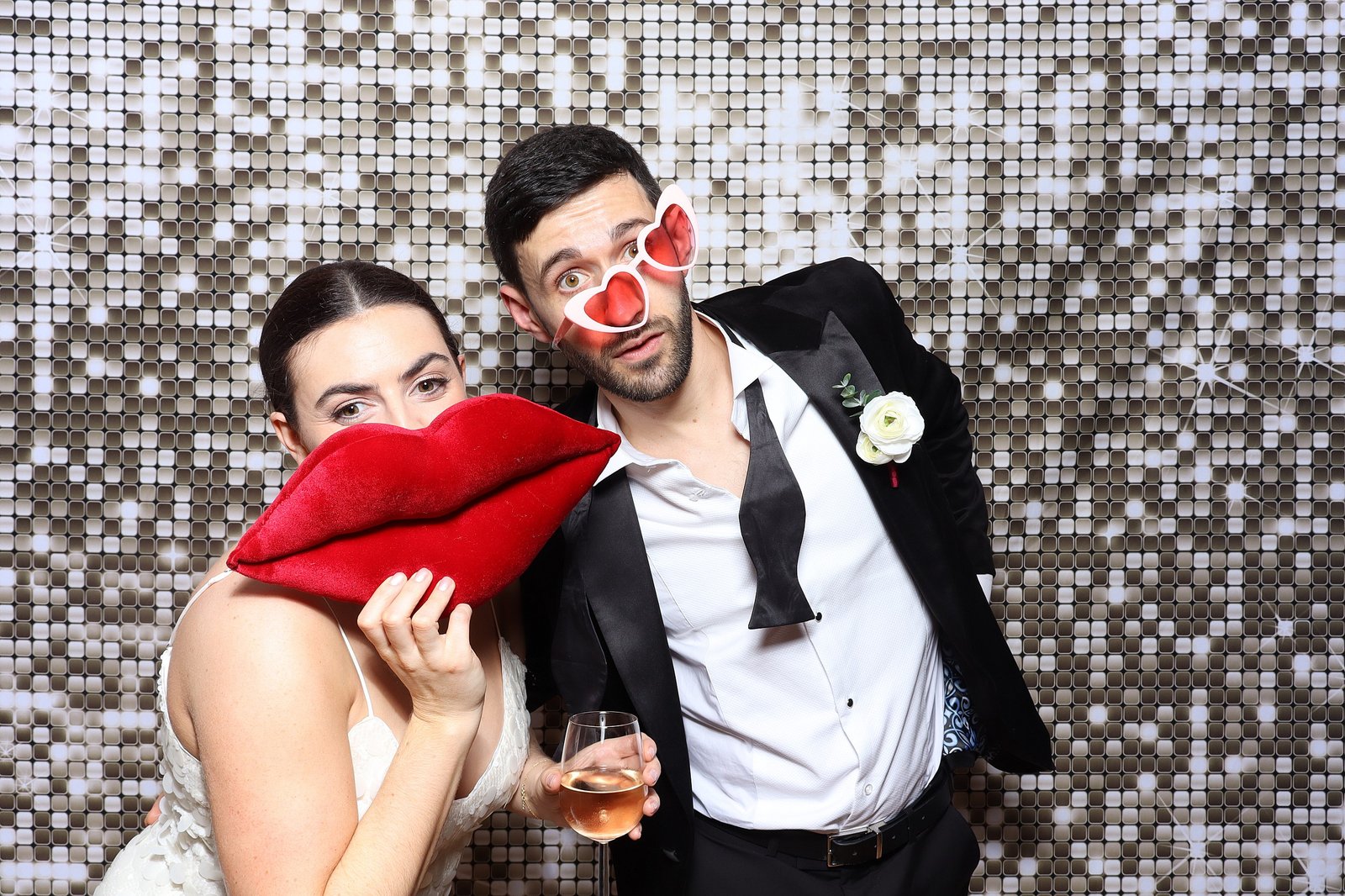 A woman holding oversized red lips and a man in heart-shaped glasses and a suit pose playfully with a drink, in front of a shimmering silver sequin backdrop—perfect for capturing fun moments with Draw Me Bot or Sketch Bot Hire.