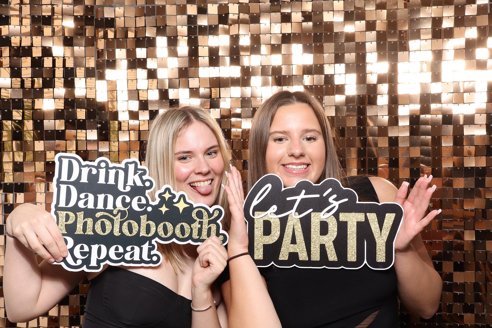 Two young women smile and pose in front of a shiny gold sequin backdrop, holding playful signs that read “Drink. Dance. Photobooth. Repeat.” and “let’s PARTY,” perfect for a corporate event experiential or graffiti wall hire experience.