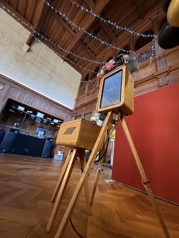 A vintage wooden television camera on a tripod stands in a large room with wood panelling, fairy lights overhead, a red backdrop, black balloons, and subtle touches reminiscent of corporate event experiential decor.