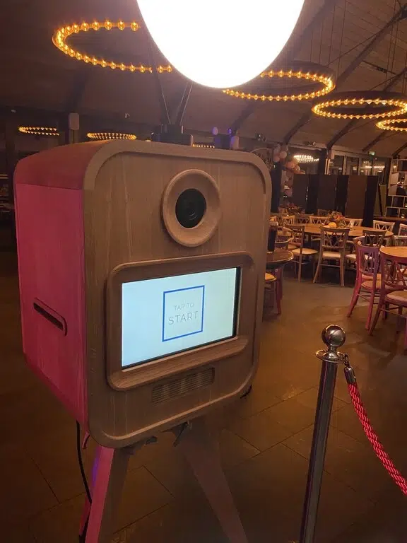 A premium photo booth with a large round light on top and a screen displaying "TAP TO START" stands in a warmly lit event space, perfect for corporate event experiential moments, with tables and chairs in the background.