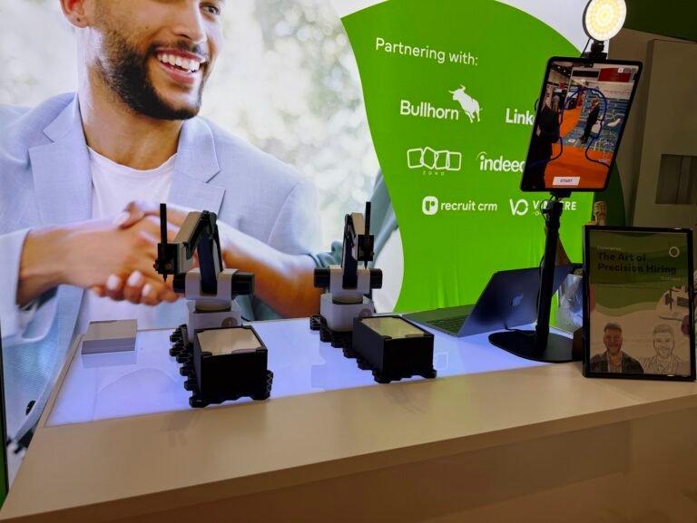 A brightly lit exhibition stand displays two robotic arms on a counter, showcasing SketchBot Activation. A large screen in the background shows a man shaking hands, with recruitment platform logos, tablets, and a framed brochure completing this Experiential setup.
