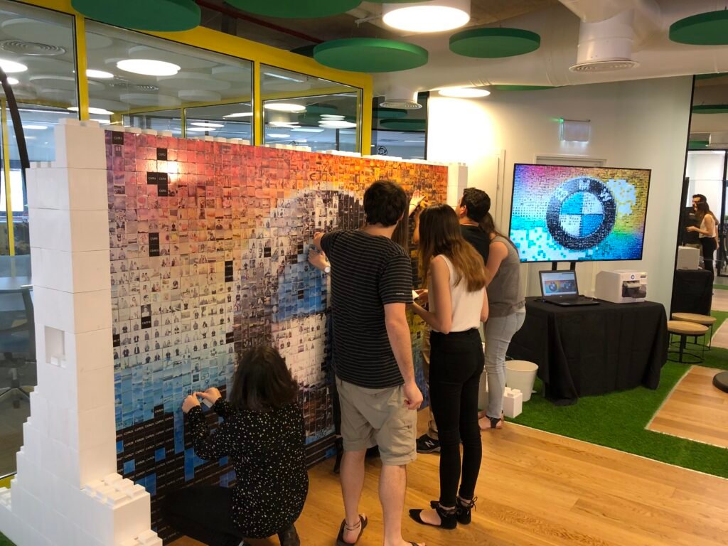 Five people work together adding tiles to a large mosaic wall featuring the BMW logo in a modern, brightly lit office with green ceiling accents—creating an engaging corporate event experiential setting. A TV displays a similar image in the background.