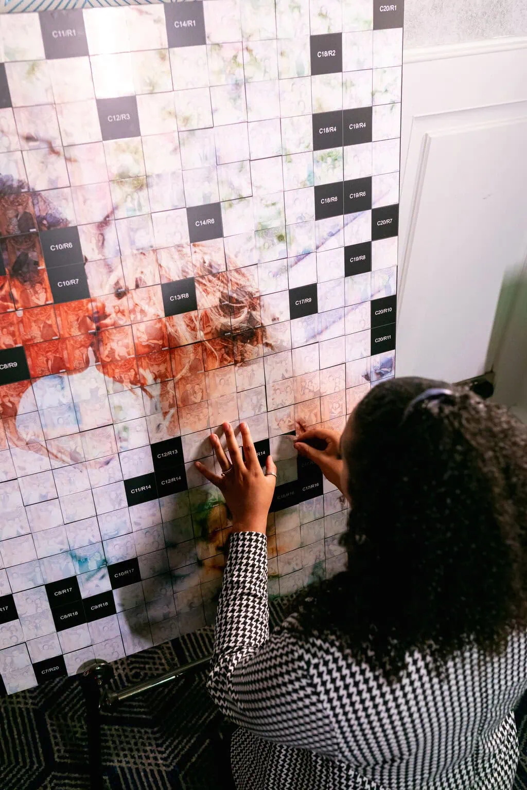 A person with curly hair places a square tile onto a large grid filled with numbered squares and partially revealed colourful image sections, enhancing a memorable corporate event experiential activity.