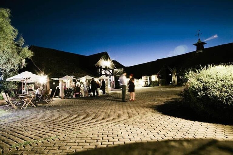 A nighttime outdoor event at Old Luxters Barn, with groups socialising under umbrellas and lights, set against a clear dark blue sky and rustic cobblestone paving in the foreground.