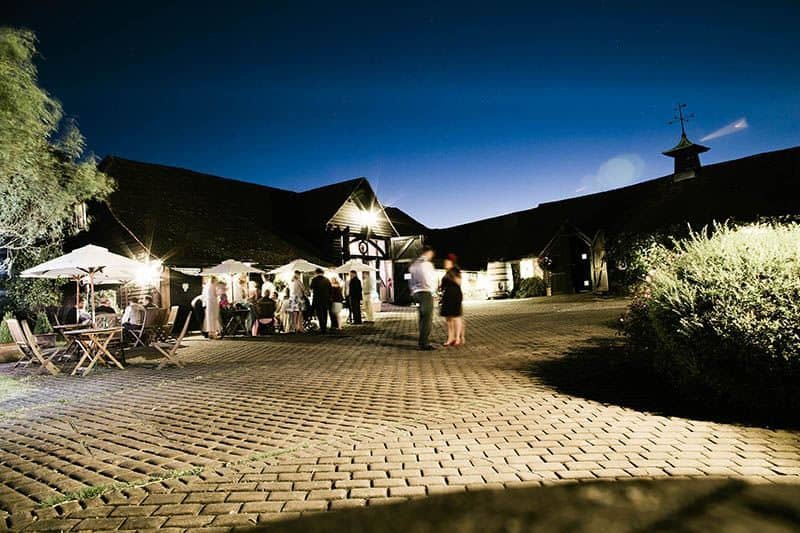 A nighttime outdoor event at Old Luxters Barn, with groups socialising under umbrellas and lights, set against a clear dark blue sky and rustic cobblestone paving in the foreground.